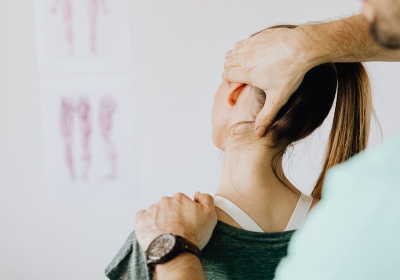 Can chiropractic help with anxiety and depression? Image shows back view of crop faceless bearded chiropractor in wristwatch checking up neck of woman while standing in front of wall with paper drawings representing body anatomy